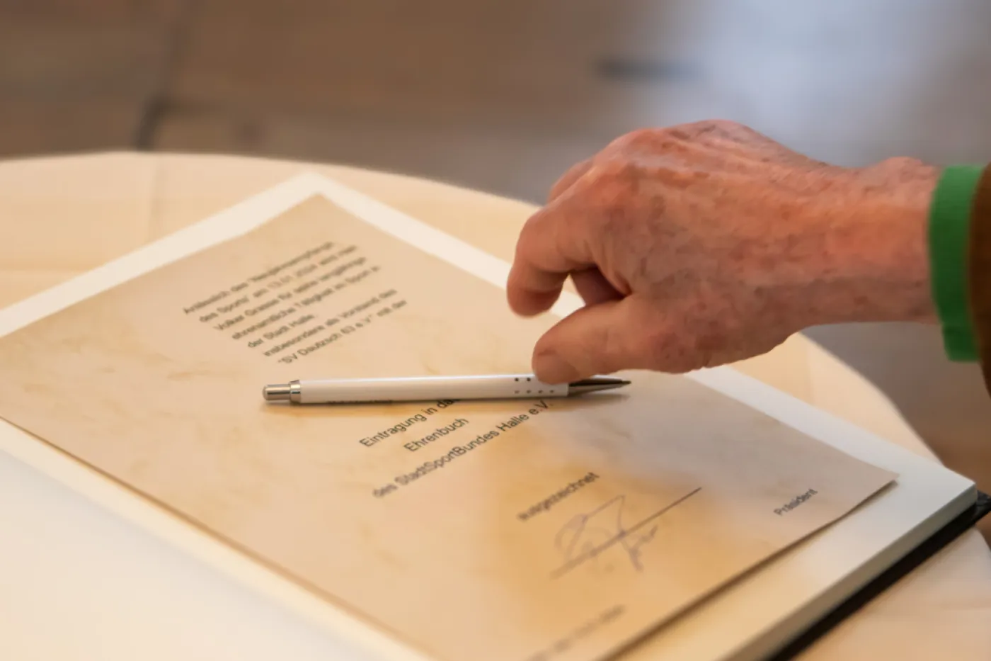 Ehrenbuch SSB Ein Buch ist aufgeschlagen, eine Urkunde klebt darin. Eine Hand greift nach dem Stift auf der Urkunde.